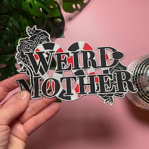 Może przedstawiać: Haftowana naszywka z napisem "WEIRD MOTHER" w kolorze czarnym, białym i czerwonym. Naszywka posiada kwiatowy wzór i unikalną czcionkę. Naszywka jest trzymana w dłoni na różowym tle.