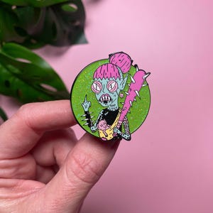 May include: A colorful enamel pin featuring a cartoonish, green-skinned creature with pink hair and a spiked club. The creature is giving the middle finger and has the text "NAP LIFE!!" on its chest. The pin is set against a glittery green circle.