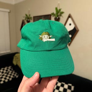 May include: A green baseball cap with a curved brim. The cap features an embroidered design of a cartoon character with a white object in its mouth. The cap is made of a durable fabric and has a classic, unstructured fit.