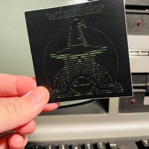 May include: A black square sticker with the word "webmaster" in green text at the top. Below the text is a pixelated image of a person wearing a hat. The sticker is held in front of a keyboard.