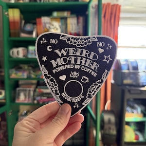 Może przedstawiać: Czarna i biała naszywka w kształcie planszy Ouija z napisem "WEIRD MOTHER POWERED BY COFFEE NEVER SLEEPS". Naszywka posiada haftowane detale, w tym gwiazdy, księżyce i kwiatowe wzory. Naszywka jest trzymana w dłoni.