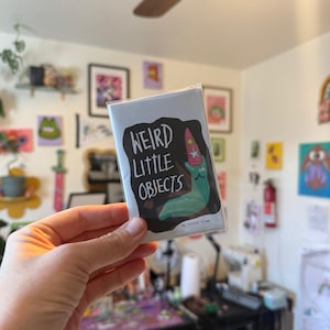 May include: A small, rectangular card with the text "WEIRD LITTLE OBJECTS" in white on a black background. The card features a whimsical illustration of a green creature with a party hat. The card is held in a person's hand.