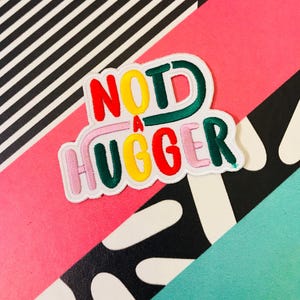 May include: A white patch with the words "NOT A HUGGER" in colorful, outlined letters. The letters are in different colors: red, yellow, green, and pink.