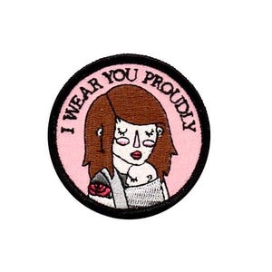 Może przedstawiać: Okrągła, haftowana naszywka z napisem "I WEAR YOU PROUDLY" na obrzeżu. Naszywka przedstawia kreskówkową ilustrację osoby z brązowymi włosami trzymającej dziecko. Tło jest różowe.