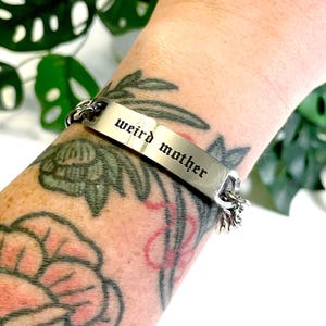 May include: A silver bracelet with a chain link design and a rectangular tag that reads "weird mother" in a gothic font.