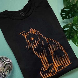 May include: A black t-shirt featuring an orange graphic of a cat silhouette composed of various computer code symbols and characters. The text cat cat.txt is printed in orange near the top of the graphic design on the front of the shirt.