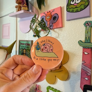May include: A round, peach-colored button with the text "she/they" and "in a little guy way." The button features a cartoon snail wearing a wizard hat. The snail has a pink shell with hearts.