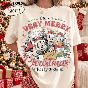 May include: An ivory short-sleeve t-shirt featuring a graphic print of Mickey Mouse, Minnie Mouse, Goofy, Donald Duck, Daisy Duck, and Pluto wearing Santa hats. The text reads Mickey's VERY MERRY Christmas Party 2026 with holiday icons.
