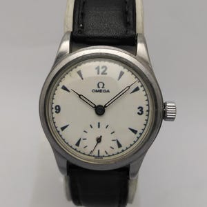 May include: An Omega wristwatch with a round silver-tone metal case and a cream-colored dial. The dial features black Arabic numerals at 3, 9, and 12, a sub-dial for seconds, and black hands. It is attached to a black leather strap with white stitching.