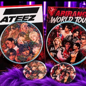 Pode incluir: Dois cartões retangulares e dois pins circulares com o grupo de K-pop ATEEZ. Um cartão exibe o texto ATEEZ, enquanto o outro diz ARIRANG WORLD TOUR. Os itens circulares e um cartão possuem um acabamento holográfico com estrelas.