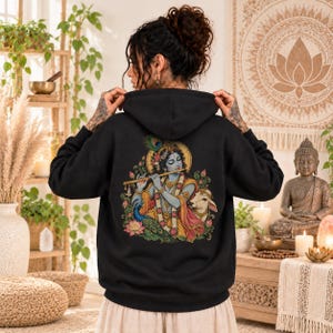 May include: A black hooded sweatshirt featuring a colorful graphic on the back. The illustration depicts a blue-skinned deity playing a golden flute, adorned with jewelry and a peacock feather, accompanied by a cow and surrounded by lotus flowers.