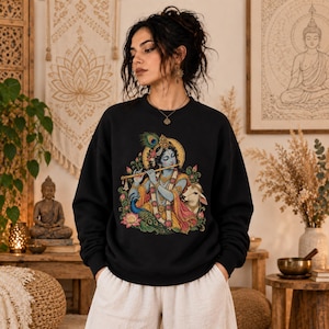 May include: A black long-sleeve sweatshirt featuring a colorful graphic print of the Hindu deity Krishna playing a flute, adorned with a peacock feather, surrounded by lotus flowers and a cow, set against a circular golden halo background.