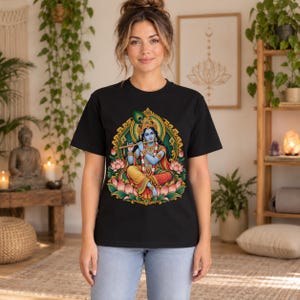 May include: A black short-sleeve t-shirt featuring a colorful graphic print of the Hindu deity Krishna playing a flute. The deity is depicted with blue skin, wearing traditional attire and jewelry, seated amidst pink lotus flowers and gold.