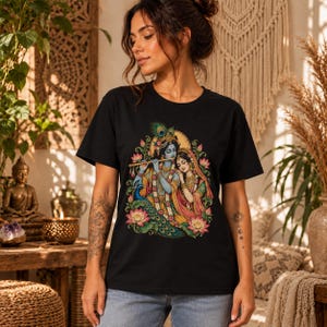 May include: A black short-sleeved t-shirt featuring a colourful graphic print of the deities Krishna and Radha. The illustration depicts Krishna playing a flute with a peacock feather in their hair, standing beside Radha, surrounded by lotus flowers.