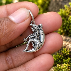 May include: A silver-toned metal pendant featuring a fairy figure with wings sitting on a crescent moon. The fairy is holding a small star-shaped object. The pendant includes a small circular jump ring at the top for attachment to a chain or cord.