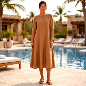 May include: A long-sleeved, calf-length dress in a solid tan color. The garment features a simple round neckline, a relaxed fit through the bodice, and a slightly flared skirt that falls below the knees. The fabric has a matte, textured appearance.