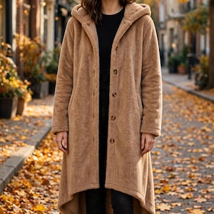 May include: A tan, knee-length, hooded coat made of a soft, fuzzy material. The coat features a front button closure with four visible brown buttons, long sleeves, and a high-low hemline. It is worn over a black shirt and black pants.
