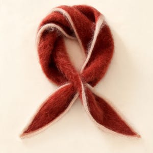 May include: A fuzzy, rust-colored scarf with a soft, brushed texture and a light beige trim along the edges. The scarf is tied in a simple loop, creating a rounded shape with two pointed ends extending downward, showcasing a distinct contrast.