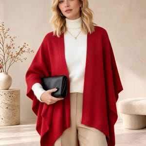 May include: A bright red open-front poncho with a ribbed hem, worn over a white long-sleeve turtleneck sweater and beige trousers. The garment features a loose, draped silhouette with wide arm openings and a smooth, solid-colored knit texture.