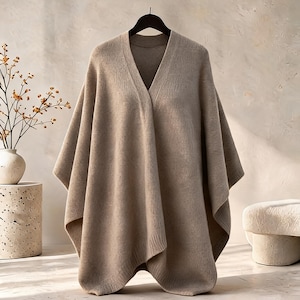 May include: A beige knit poncho featuring a V-neckline and a relaxed, draped silhouette. The garment is constructed from a soft, textured fabric with ribbed detailing along the edges and neckline, hanging from a black hanger.