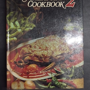 Könnte beinhalten: Ein gebundenes Buch mit dem Titel The Malaysian Cookbook 2 vor dunklem Hintergrund. Das Cover zeigt ein Foto eines gekochten Gerichts auf einem weißen Teller, umgeben von frischem grünen Gemüse, roten Chilischoten und Zwiebeln auf dunklem Grund.
