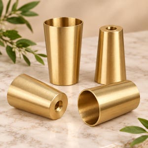 Könnte beinhalten: Vier konische, zylindrische Objekte aus gebürstetem goldfarbenem Metall. Zwei der Objekte stehen aufrecht, während zwei auf der Seite liegen. Jedes Teil hat eine glatte, metallische Oberfläche und eine hohle Mitte mit einer Gewindeöffnung.