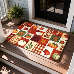 Peut inclure: Un paillasson rectangulaire présentant un motif en patchwork de carrés sur le thème de l'automne. Le design inclut des citrouilles orange, blanches et vertes, des feuilles d'érable rouges et orange, ainsi que des fonds à carreaux ou à pois.