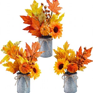 May include: Three decorative arrangements featuring artificial autumn foliage, orange pumpkins, sunflowers, and brown berries. Each bouquet is housed in a cylindrical, corrugated galvanized metal container tied with a rustic jute twine bow.
