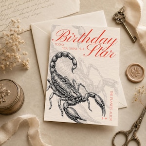 May include: A rectangular greeting card featuring a black ink illustration of a scorpion. The text reads Birthday Star, Today, You Shine, and The Planets Align in red script and serif fonts. The card is placed on a light-coloured surface.