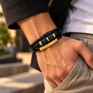 May include: A multi-strand bracelet featuring black braided leather cords and smooth black leather straps. The design includes gold-tone metal accents, a rectangular gold-tone plate, and a black cylindrical clasp with gold-tone rings.