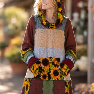 Puede incluir: Una túnica con capucha y mangas largas que presenta un diseño de patchwork con tela de girasoles en la capucha, los puños y el bolsillo delantero. La prenda incluye paneles de tejido de punto trenzado en beige, azul claro y rojo oscuro.