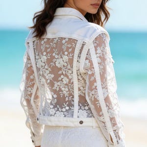 May include: White denim jacket with a floral lace back panel. The jacket has long sleeves, a button-down front, and a matching white belt. The model is wearing a white lace skirt. The background is a beach with blue water.