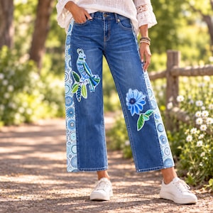 May include: Dark blue denim jeans featuring wide side panels with a light blue and white circular mandala pattern. The front of the jeans displays embroidered designs of a blue bird perched on green leaves and a large blue flower with green leaves.