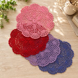 May include: A set of four round, circular crochet doilies featuring intricate openwork patterns. The doilies are colored in shades of pink, red, light purple, and dark blue, each with a scalloped edge and a central circular motif design.