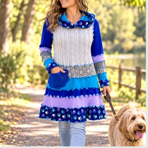 May include: A long-sleeved tunic featuring a grey cable-knit bodice, a cowl neck, and a tiered skirt with blue, purple, and grey sections. The garment includes a blue lace pocket detail and a pattern of colorful paw prints on the hem and collar.