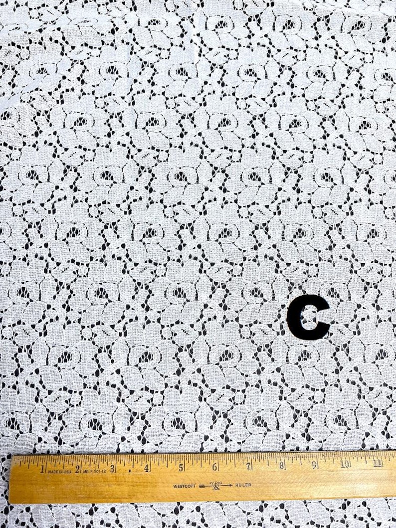 May include: White lace fabric with a floral pattern. A wooden ruler with inch markings is at the bottom. A black letter "C" is in the center. The fabric has a delicate, open weave design.