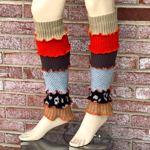 May include: A pair of vibrant leg warmers on a mannequin. The leg warmers are multi-layered, showcasing tan, red, brown, light blue, and black with a spotted pattern. Each layer is trimmed with a ruffled edge, adding a decorative touch.