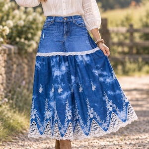 Könnte beinhalten: Ein langer Jeansrock in mittlerer Waschung mit einem abgenutzten oberen Bereich. Der untere Teil zeigt einen Batik-Effekt in Blau- und Weißtönen, akzentuiert durch weiße Blumenstickereien und einen breiten weißen Spitzenbesatz am Saum.