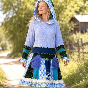 May include: A hooded dress featuring a light blue cable-knit bodice and a multi-patterned skirt. The hood and skirt display a blue and white gingham print with white daisy motifs. The skirt includes dark green plaid panels and purple crochet pockets.