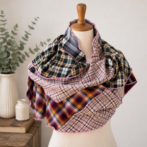 May include: A multi-layered infinity scarf featuring a patchwork of various plaid patterns in shades of forest green, mustard yellow, burgundy, black, white, and soft pink, finished with a delicate light pink stitched edge throughout the fabric.