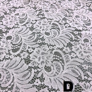 May include: Close-up of white lace fabric with a floral pattern. The delicate lace features intricate designs and a textured surface. The letter "D" is visible in the lower right corner. This fabric is suitable for various craft and sewing projects.