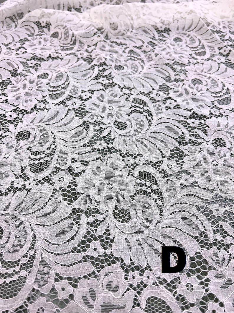 May include: Close-up of white lace fabric with a floral pattern. The delicate lace features intricate designs and a textured surface. The letter "D" is visible in the lower right corner. This fabric is suitable for various craft and sewing projects.
