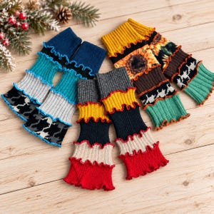 May include: Three pairs of knit leg warmers featuring ruffled edges. One pair is blue, gray, and black with a houndstooth pattern. Another pair features yellow, black, and red stripes. The third pair displays a sunflower print with green accents.