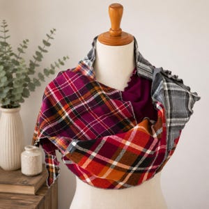 May include: A multi-patterned infinity scarf featuring a mix of red, orange, and black plaid fabric panels combined with solid deep magenta sections and a grey and white plaid accent, draped over a wooden mannequin torso for display.