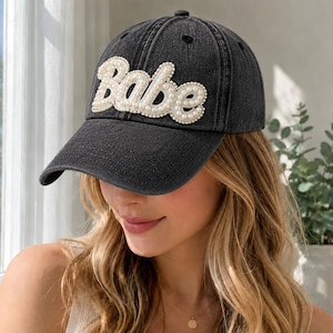 May include: A charcoal grey denim baseball cap featuring the word Babe in white, raised, pearl-like embellishments across the front panel. The cap has a structured crown, a curved brim, and stitched eyelets on the top panels for ventilation.