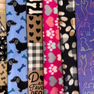 May include: A collection of colorful fleece fabrics with various dog-themed patterns. Designs include dachshunds, bones, hearts, paw prints, and plaid. The fabrics are in shades of blue, pink, black, brown, and purple, suitable for pet accessories.
