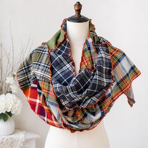 May include: A multi-colored infinity scarf featuring a patchwork of various plaid patterns in shades of olive green, navy blue, red, and white. The scarf has a ruffled edge finished with a bright orange stitched trim, creating a layered look.