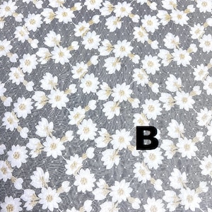 May include: White floral lace fabric with a gray background. The fabric features a repeating pattern of white flowers with gold accents. A wooden ruler is at the bottom of the image, with the letter "B" in black.