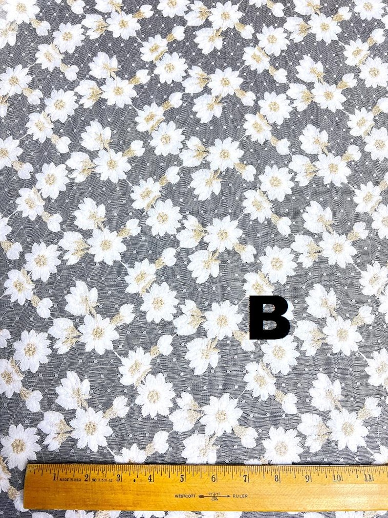 May include: White floral lace fabric with a gray background. The fabric features a repeating pattern of white flowers with gold accents. A wooden ruler is at the bottom of the image, with the letter "B" in black.