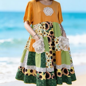 Könnte beinhalten: Ein Patchwork-Kleid mit Sonnenblumenmuster, grünen und orangefarbenen Einsätzen. Das Kleid hat kurze Ärmel, einen runden Ausschnitt und mit Spitze besetzte Taschen. Eine weiße Spitzenapplikation ziert das Oberteil. Das Kleid ist knielang.